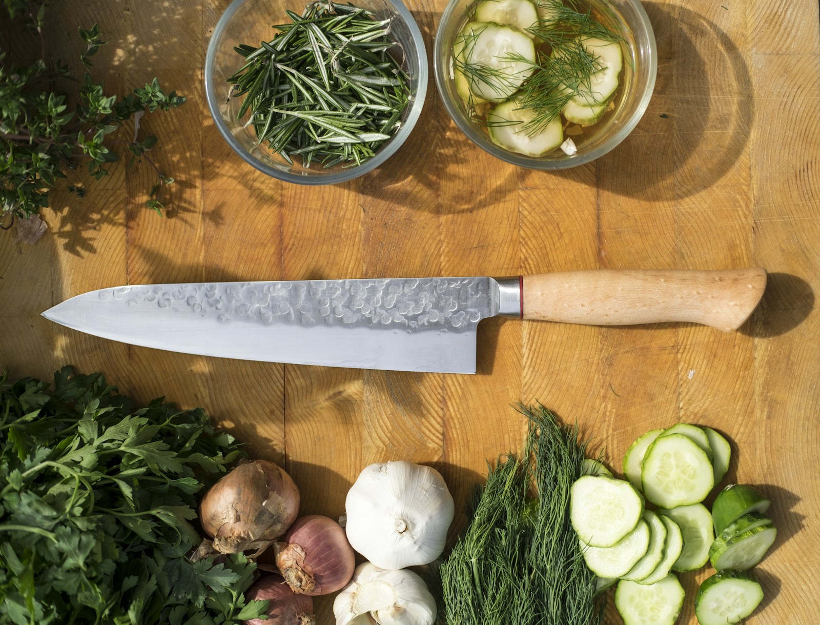 A long, pointed kitchen knife on a wooden board with sliced cucumber, herbs, and other vegetables.